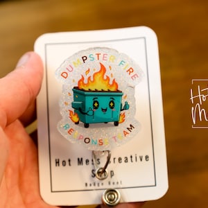Puede incluir: Un portatarjetas con un diseño de dibujos animados de un contenedor de basura en llamas. El contenedor de basura turquesa tiene una cara sonriente y un pulgar hacia arriba. El texto dice "Dumpster Fire Response Team". El portatarjetas está en una tarjeta blanca con el texto "Hot Mess Creative Shop".