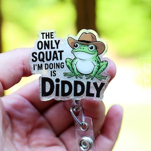 May include: A clear acrylic badge reel with a cartoon frog wearing a cowboy hat. The text reads "THE ONLY SQUAT I'M DOING IS DIDDLY" in black lettering. The frog is green and white.