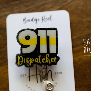 Puede incluir: Un portatarjetas con un dise&ntilde;o 911 negro y brillante, con una franja amarilla y la palabra "Dispatcher". El portatarjetas est&aacute; sujeto a una tarjeta blanca con el texto "Badge Reel" y "Hot Mess Creative Shop".
