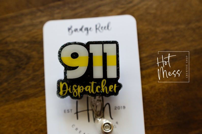 Puede incluir: Un portatarjetas con un dise&ntilde;o 911 negro y brillante, con una franja amarilla y la palabra "Dispatcher". El portatarjetas est&aacute; sujeto a una tarjeta blanca con el texto "Badge Reel" y "Hot Mess Creative Shop".