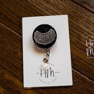 May include: A black retractable badge holder with a white outline of a judge's collar. The badge holder is on a white card with the text "Hot Mess Creative Shop" and "Hm Est 2019 Creative Shop".