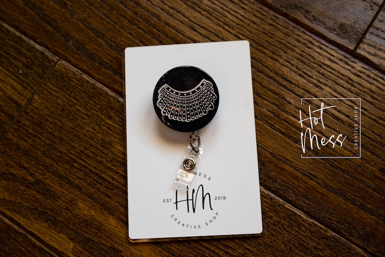 May include: A black retractable badge holder with a white outline of a judge's collar. The badge holder is on a white card with the text "Hot Mess Creative Shop" and "Hm Est 2019 Creative Shop".