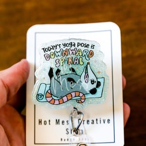Op de afbeelding: Een heldere, glinsterende badge met een cartoon opossum in een yogahouding op een blauwe mat. De tekst luidt "Today's yoga pose is DOWNWARD SPIRAL". De badge heeft een zilveren clip en de tekst "Hot Mess Creative Shop".