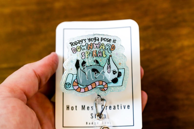 Op de afbeelding: Een heldere, glinsterende badge met een cartoon opossum in een yogahouding op een blauwe mat. De tekst luidt "Today's yoga pose is DOWNWARD SPIRAL". De badge heeft een zilveren clip en de tekst "Hot Mess Creative Shop".