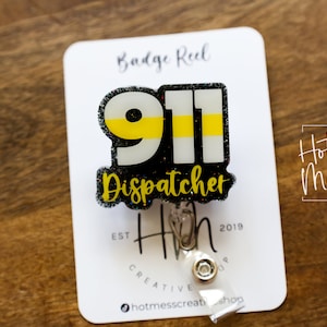 May include: A badge reel featuring a 911 Dispatcher design. The design includes the number 911 in white and yellow, with the word "Dispatcher" in yellow script. The badge reel is attached to a white card with the "Hot Mess Creative Shop" logo.