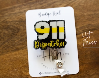 Op de afbeelding: Een badge reel met een 911 Dispatcher ontwerp. Het ontwerp toont het nummer 911 in wit en geel, met het woord "Dispatcher" in gele letters. De badge reel is bevestigd aan een witte kaart met het logo "Hot Mess Creative Shop".