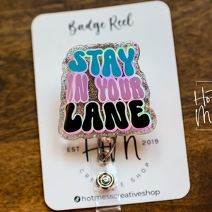 May include: A silver glitter badge reel with a colourful, glittery,  "Stay in your lane" design. The badge reel is attached to a white card with the text "Hot Mess Creative Shop" and "Est. 2019".