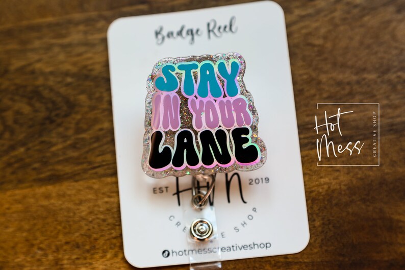 May include: A silver glitter badge reel with a colourful, glittery,  "Stay in your lane" design. The badge reel is attached to a white card with the text "Hot Mess Creative Shop" and "Est. 2019".