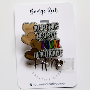 May include: A silver glitter badge reel with a clear plastic badge holder. The reel features a design of four brown hearts with the text "All Bodies Deserve Equal Healthcare" in rainbow colors. The text "Hot Mess Creative Shop" is printed on the card.