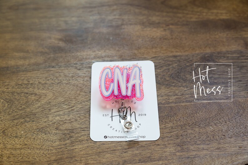 K&ouml;nnte beinhalten: Rosa-wei&szlig;er Glitzer-CNA-Badge-Rollenhalter mit einem silbernen einziehbaren Badge-Halter. Der Badge-Rollenhalter tr&auml;gt den Text "Hot Mess Creative Shop" und "Est 2019".