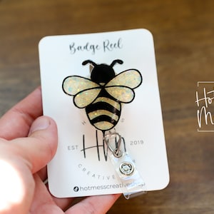 May include: A bee-shaped badge reel with a black and yellow design, featuring glittery wings. The badge reel is attached to a white card with the text "Badge Reel" and "Hot Mess Creative Shop".