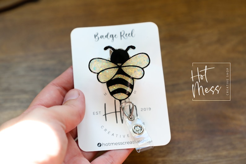 May include: A bee-shaped badge reel with a black and yellow design, featuring glittery wings. The badge reel is attached to a white card with the text "Badge Reel" and "Hot Mess Creative Shop".