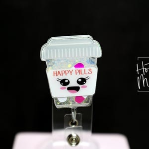May include: A white and clear badge reel featuring a cartoon coffee cup design. The cup has a smiling face and the words "HAPPY PILLS" in pink. The cup is filled with glitter and has a clear lid. The badge reel is attached to a clear plastic clip.