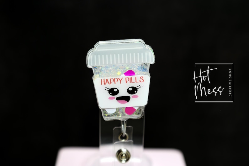 May include: A white and clear badge reel featuring a cartoon coffee cup design. The cup has a smiling face and the words "HAPPY PILLS" in pink. The cup is filled with glitter and has a clear lid. The badge reel is attached to a clear plastic clip.