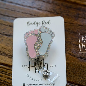 May include: A silver badge reel with a pink and blue glitter baby footprint charm. The charm is attached to a clear plastic retractable cord. The card reads "Badge Reel" and "Hot Mess Creative Shop".