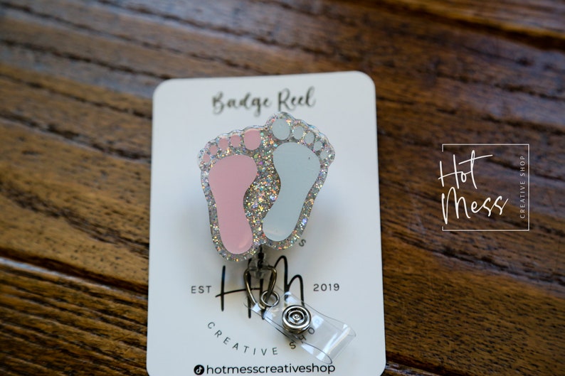 May include: A silver badge reel with a pink and blue glitter baby footprint charm. The charm is attached to a clear plastic retractable cord. The card reads "Badge Reel" and "Hot Mess Creative Shop".