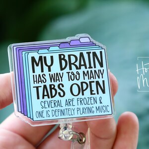 May include: A clear, rectangular badge reel with a design featuring the text "MY BRAIN HAS WAY TOO MANY TABS OPEN." The design includes purple and blue tabs. The words "SEVERAL ARE FROZEN & ONE IS DEFINITELY PLAYING MUSIC" are also included.