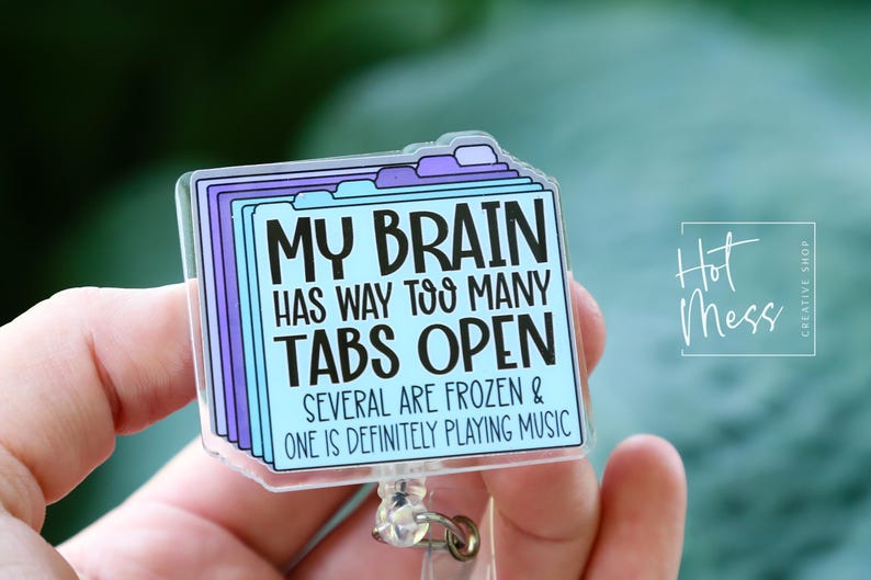 May include: A clear, rectangular badge reel with a design featuring the text "MY BRAIN HAS WAY TOO MANY TABS OPEN." The design includes purple and blue tabs. The words "SEVERAL ARE FROZEN & ONE IS DEFINITELY PLAYING MUSIC" are also included.