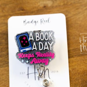 May include: A silver glitter badge reel with a blue book and pink text that reads "A Book A Day Keeps Reality Away".