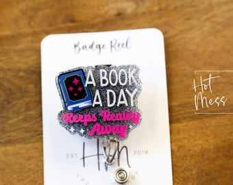 Puede incluir: Un carrete de insignia de purpurina plateada con un libro azul y texto rosa que dice "A Book A Day Keeps Reality Away".