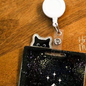 Puede incluir: Un dije de carrete de insignia de gato negro y brillante que asoma por encima de un porta-insignias negro y colorido. El porta-insignias tiene un diseño de galaxia con estrellas blancas y motas de colores. El carrete es blanco.