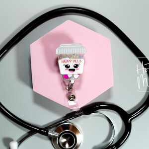 May include: A novelty badge reel featuring a cartoon pill bottle design with the words "Happy Pills". The reel is set against a pink hexagonal backdrop, with a black stethoscope in the foreground. The image also includes the text "Hot Mess Creative Shop."