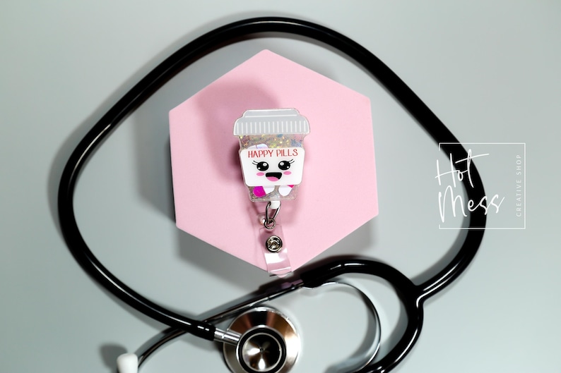 May include: A novelty badge reel featuring a cartoon pill bottle design with the words "Happy Pills". The reel is set against a pink hexagonal backdrop, with a black stethoscope in the foreground. The image also includes the text "Hot Mess Creative Shop."