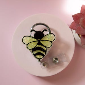 May include: A bee-shaped badge reel with a black and yellow design, featuring glittery wings. The badge reel is attached to a clear plastic strap and a metal clip. The background is a light pink color.