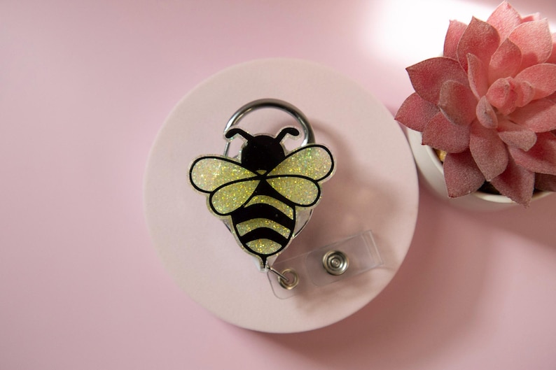 May include: A bee-shaped badge reel with a black and yellow design, featuring glittery wings. The badge reel is attached to a clear plastic strap and a metal clip. The background is a light pink color.