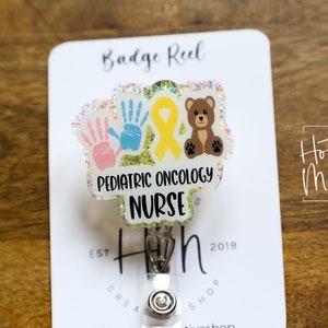 Peut inclure: Un porte-badge blanc avec un charme en plastique transparent repr&eacute;sentant un ruban jaune, deux empreintes de mains et un ours en peluche brun. Le charme porte le texte "Pediatric Oncology Nurse".