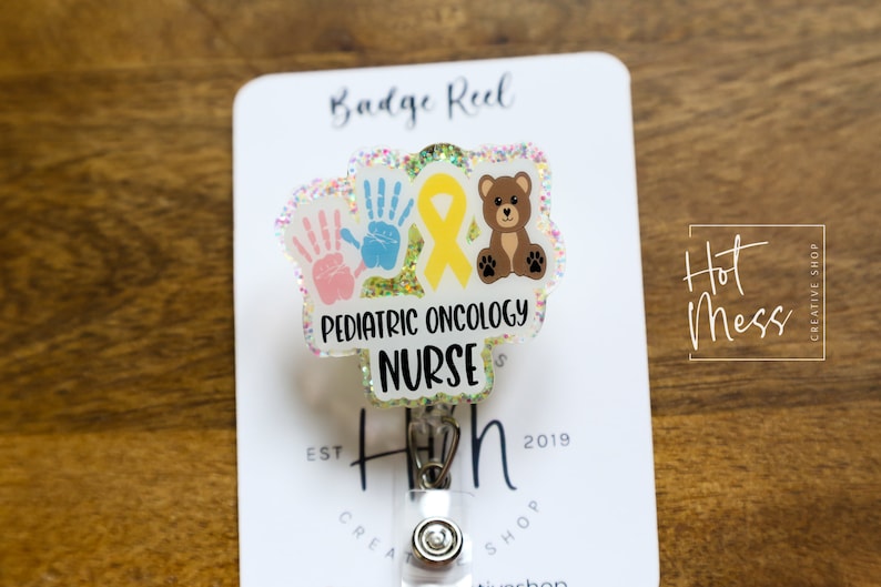 Peut inclure: Un porte-badge blanc avec un charme en plastique transparent repr&eacute;sentant un ruban jaune, deux empreintes de mains et un ours en peluche brun. Le charme porte le texte "Pediatric Oncology Nurse".