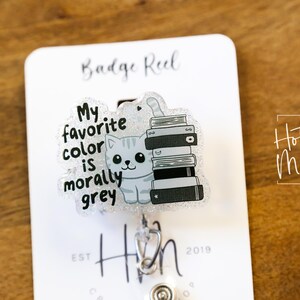May include: A badge reel with a clear plastic charm featuring a cartoon cat with a stack of books. The text on the charm reads "My favorite color is morally grey".