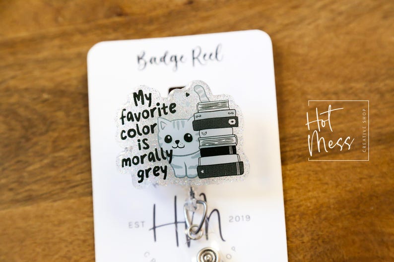 May include: A badge reel with a clear plastic charm featuring a cartoon cat with a stack of books. The text on the charm reads "My favorite color is morally grey".