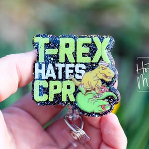 May include: A black and green glitter badge holder with a green T-Rex dinosaur graphic and the text "T-Rex Hates CPR".