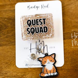 Könnte beinhalten: Goldener Glitzer-Badge-Rollenhalter mit einem schwarz-weißen "QUEST SQUAD"-Design. Der Rollenhalter hat eine transparente, einziehbare Schnur aus Kunststoff und einen niedlichen, orangefarbenen Katzen-Anhänger.