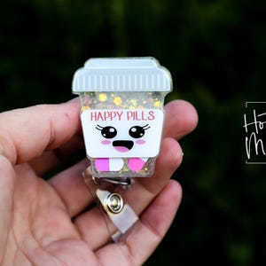 May include: A badge reel featuring a cartoon coffee cup design. The cup is white with the words "HAPPY PILLS" in pink. The cup has a smiling face and pink and white pills at the bottom. The reel is held in a hand.