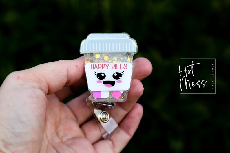 May include: A badge reel featuring a cartoon coffee cup design. The cup is white with the words "HAPPY PILLS" in pink. The cup has a smiling face and pink and white pills at the bottom. The reel is held in a hand.