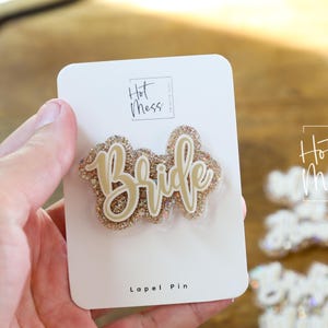 Może przedstawiać: Złoty brokatowy pin "Bride" na białej karcie. Pin ma kursywę i jest otoczony złotym brokatem. Na karcie znajduje się tekst "Hot Mess" i "Lapel Pin".