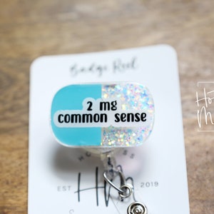 May include: A blue and white glitter pill-shaped badge reel with the text "2 mg common sense".
