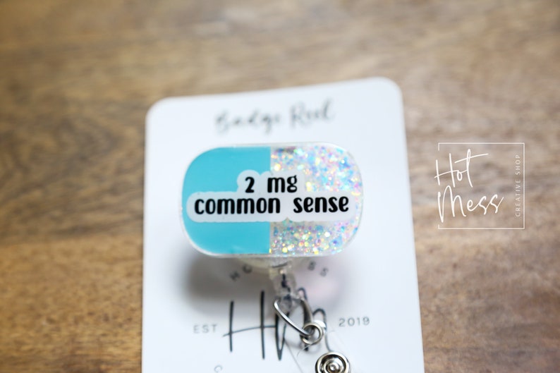 May include: A blue and white glitter pill-shaped badge reel with the text "2 mg common sense".
