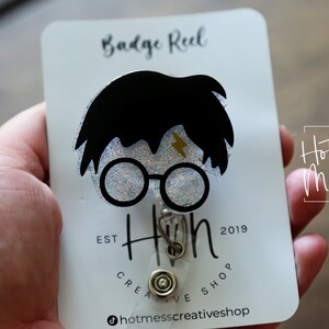 Puede incluir: Una tarjeta blanca con texto negro que dice "Badge Reel" y "Hot Mess Creative Shop" con un carrete de insignia de brillo negro y plateado con una silueta de Harry Potter con una cicatriz de rayo y gafas redondas. El texto "EST 2019" tambi&eacute;n est&aacute; en la tarjeta.