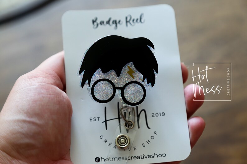 Puede incluir: Una tarjeta blanca con texto negro que dice "Badge Reel" y "Hot Mess Creative Shop" con un carrete de insignia de brillo negro y plateado con una silueta de Harry Potter con una cicatriz de rayo y gafas redondas. El texto "EST 2019" tambi&eacute;n est&aacute; en la tarjeta.