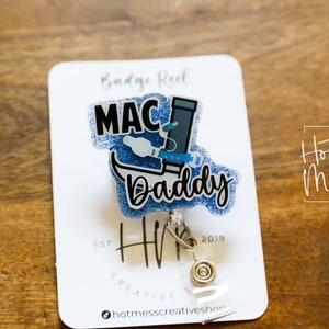 May include: Blue glitter badge reel with a white and blue illustration of a medical device and the text "MAC Daddy". The badge reel is attached to a white card with the text "Hot Mess Creative Shop" and "Badge Reel".