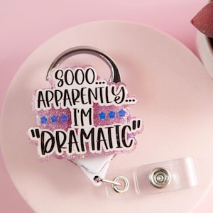 Könnte beinhalten: Ein Ausweis-Rollhalter mit einem glitzernden, rosa-weißen Design. Der Ausweis-Rollhalter hat den Text "SOOO... APPARENTLY... I'M "DRAMATIC"" in schwarzer Schrift. Die Rolle ist an einem transparenten Kunststoffclip befestigt.