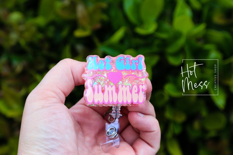 Pode incluir: Chaveiro de resina rosa com glitter com o texto "Hot Girl Summer" em letras azuis e rosas com um cora&ccedil;&atilde;o rosa no meio.
