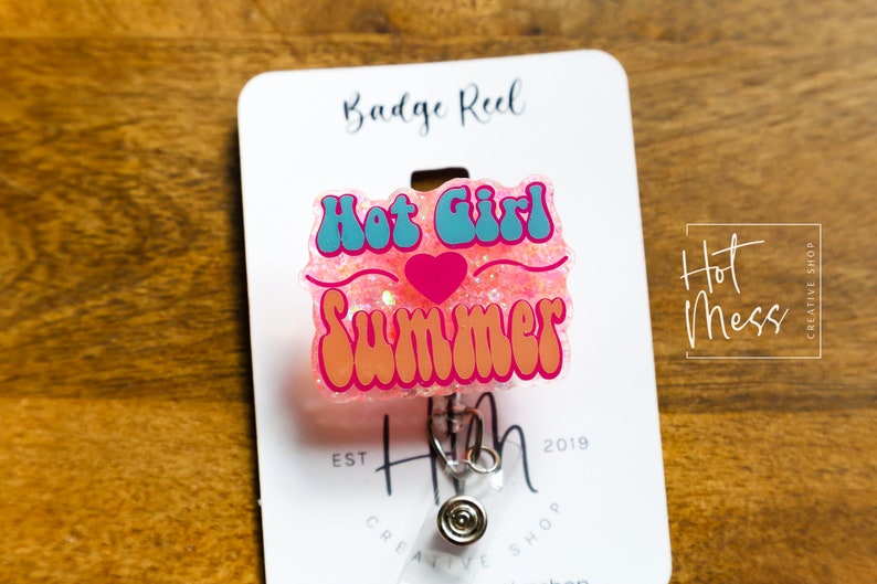 Pode incluir: Carretel de crach&aacute; de glitter rosa com um design "Hot Girl Summer" azul e rosa. O carretel &eacute; preso a um suporte de crach&aacute; retr&aacute;til prateado.