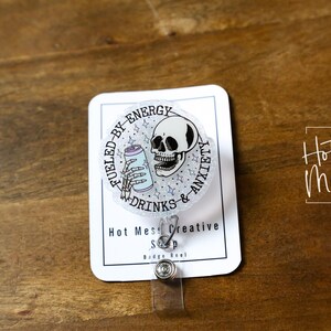 May include: A clear badge reel with a circular design featuring a skull, a can, and the text "Fueled by Energy Drinks & Anxiety." The badge reel has a clear plastic strap and a metal clip. The words "Hot Mess Creative Shop" are also visible.