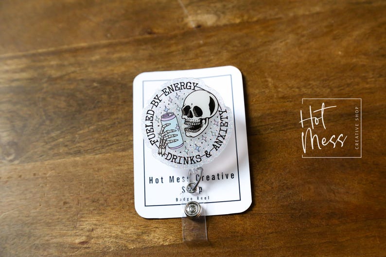 May include: A clear badge reel with a circular design featuring a skull, a can, and the text "Fueled by Energy Drinks & Anxiety." The badge reel has a clear plastic strap and a metal clip. The words "Hot Mess Creative Shop" are also visible.