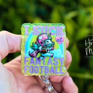 May include: A glittery yellow and green badge with a unicorn wearing a football helmet and holding a football. The badge says "I suck at Fantasy Football" with a pink heart that says "at".