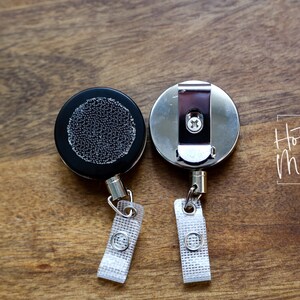 May include: Two retractable badge reels, one with a black plastic case and a textured surface, and the other with a silver metal case. Both reels have a silver metal clip and a white fabric strap.